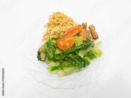 Fried noodles on a white plate topped with sliced green vegetables plus tomatoes and fried tempeh slices
