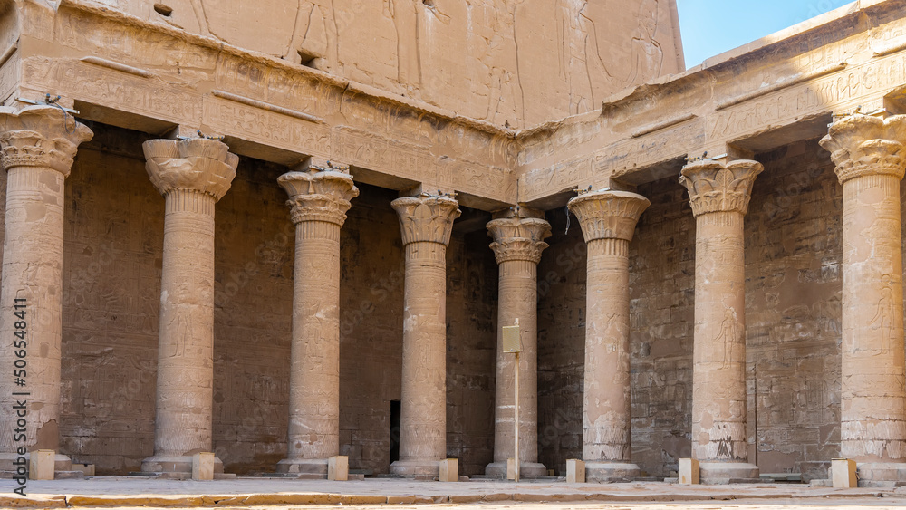 Colonnade in the Temple of Horus in Edfu. Tall columns with various carved capitals stand in a row and in the corner of the hall. Hieroglyphs, drawings of gods and pharaohs are visible. Egypt