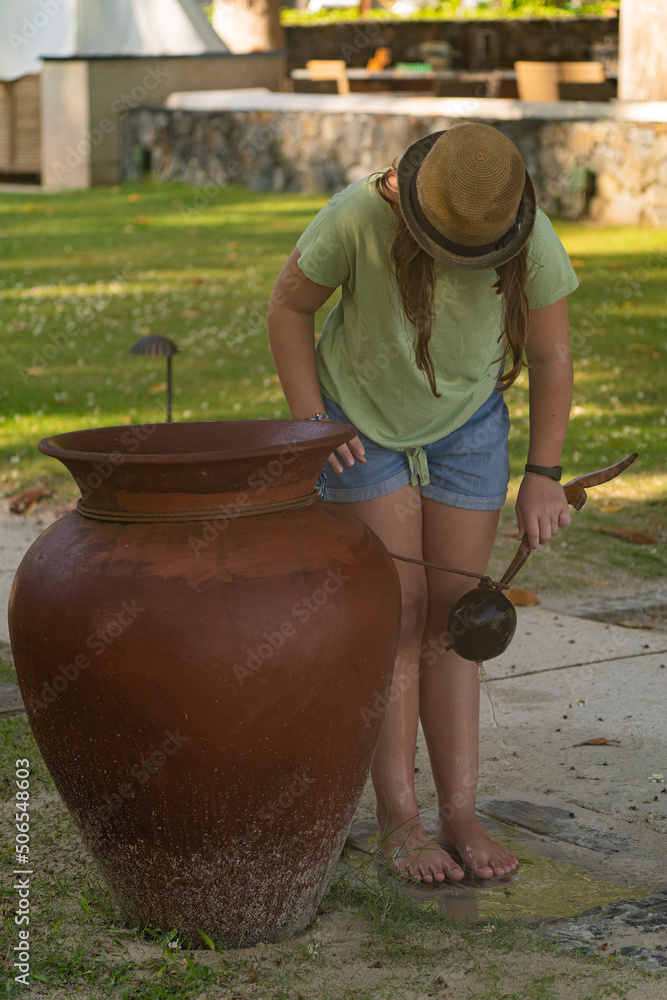 Obraz premium A girl wearing summer clothes washes her feet and legs on the beach using water from a big ceramic pot. 