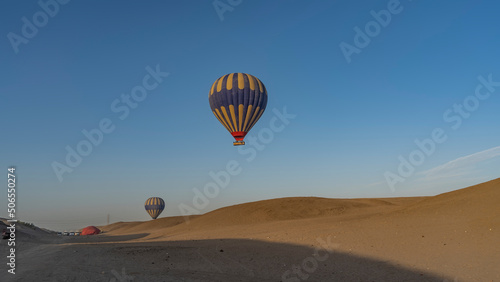 Bright balloons are flying in a clear blue sky. Below - the sand dunes of the desert. Several cars are waiting in the distance. Light and shadows. Egypt. Luxor