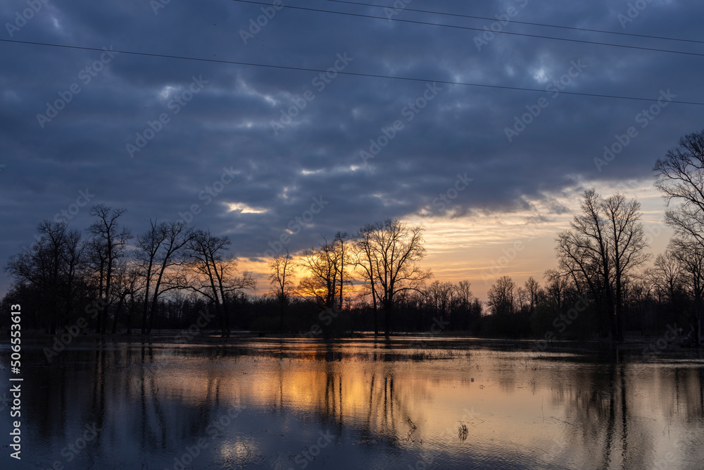 Dramatic evening landscape. The sunset sky is reflected in the water. Silhouettes of fabulous trees at sunset. The river overflowed its banks.