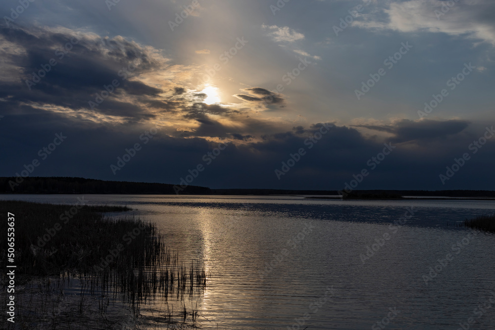 Evening dramatic landscape, lake in spring, sun shining through blue clouds. Ripples on the water from a strong wind.