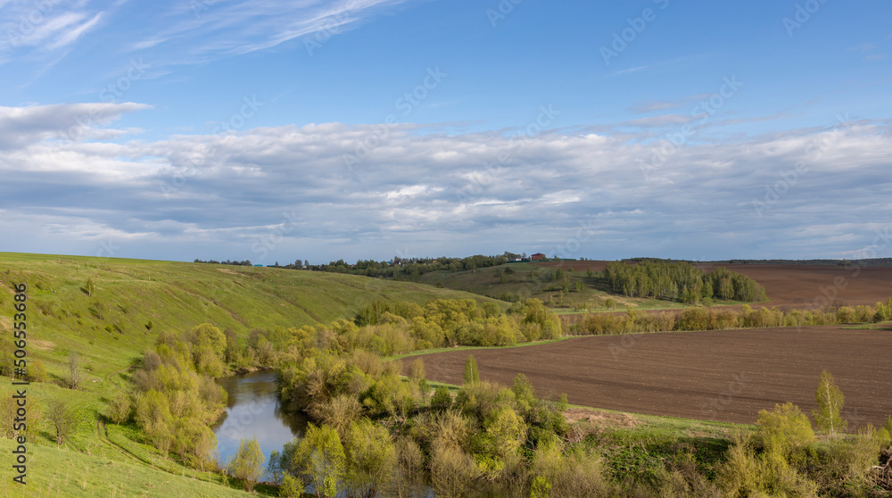 View of the countryside. bright greenery in the ravine. Saturated green grass against the blue sky. Plowed field on the horizon. Landscape with a small river in a ravine.