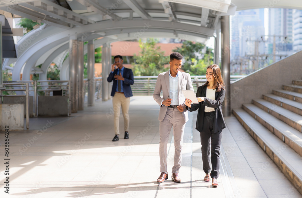 businesswoman and businessman working with tablet together outside building in city. caucasian woman and black man using tablet, executive man using mobile and calling at background