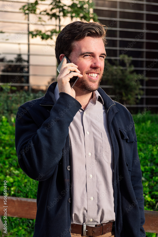 smiling caucasian business man talking by phone in a park next to an office building, concept of technology and communication smiling caucasian business man talking by phone in a park next to an office building, concept of technology and communication