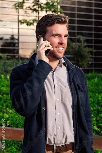 smiling caucasian business man talking by phone in a park next to an office building, concept of technology and communication