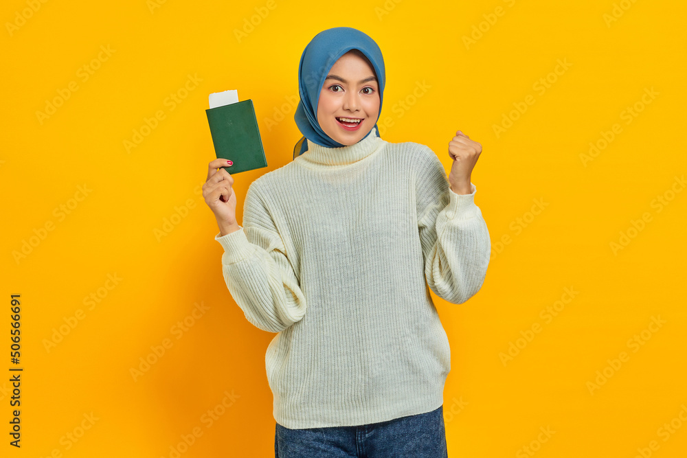 Excited beautiful Asian woman in white sweater holding passport and celebrating big luck and success with raised fists isolated over yellow background. Summer Trip Excited beautiful Asian woman in white sweater holding passport and celebrating big luck and success with raised fists isolated over yellow background. Summer Trip