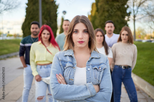 Self assured young female with crossed arms in denim jacket looking at camera while standing on street against friends on city park