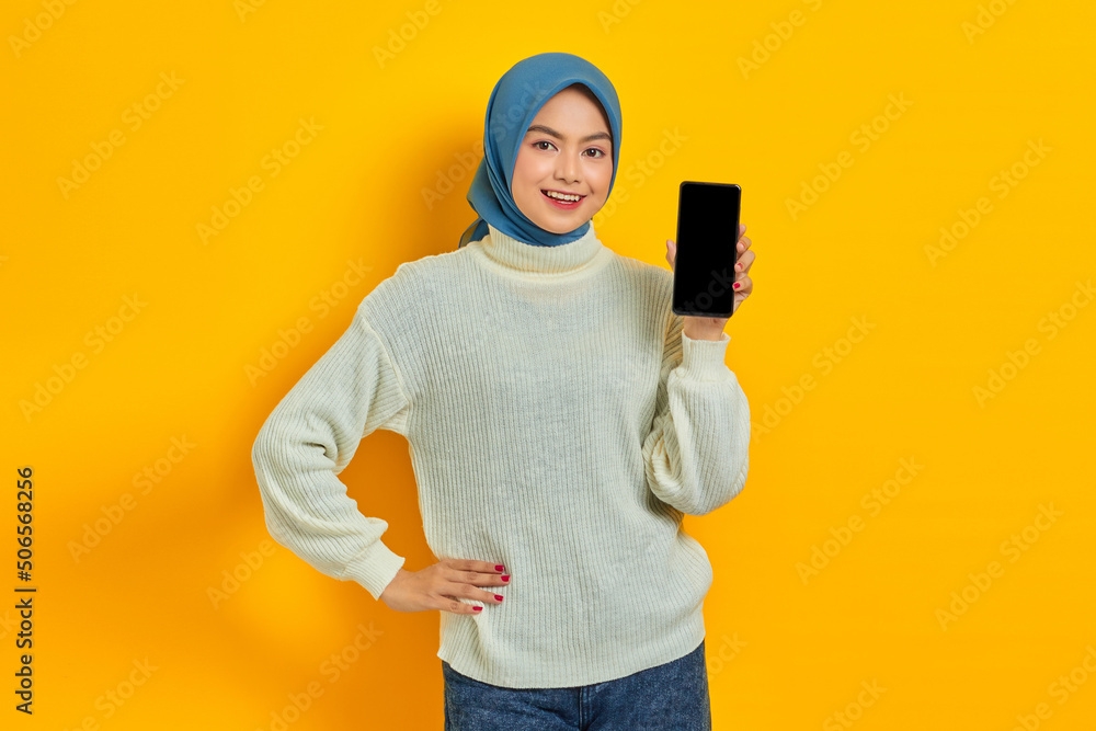 Smiling beautiful Asian woman in white sweater and hijab, showing blank mobile phone screen isolated on yellow background. Muslim lifestyle concept Smiling beautiful Asian woman in white sweater and hijab, showing blank mobile phone screen isolated on yellow background. Muslim lifestyle concept