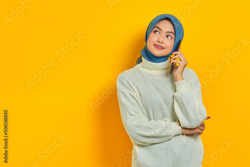 Smiling young asian woman in white sweater and hijab talking on mobile while looking at copy space isolated over yellow background. People islam religious concept © Bangun Stock Photo