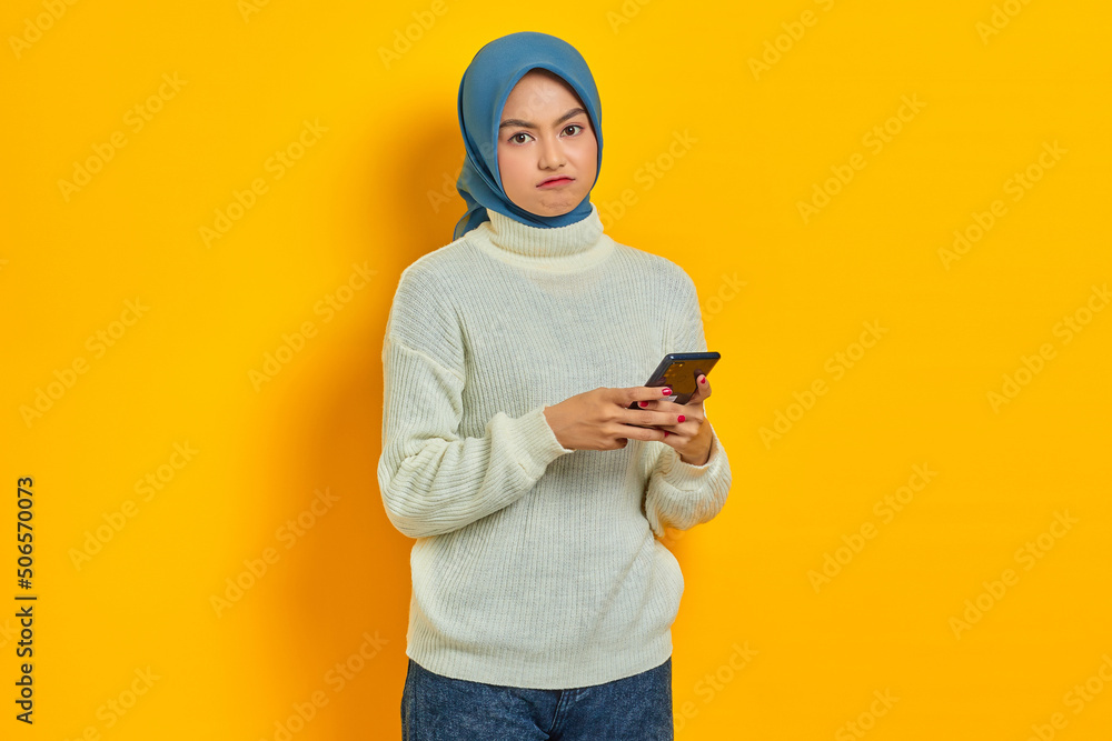 Pensive young asian woman in white sweater and hijab using on mobile phone and looking at camera isolated over yellow background. People islam religious concept Pensive young asian woman in white sweater and hijab using on mobile phone and looking at camera isolated over yellow background. People islam religious concept