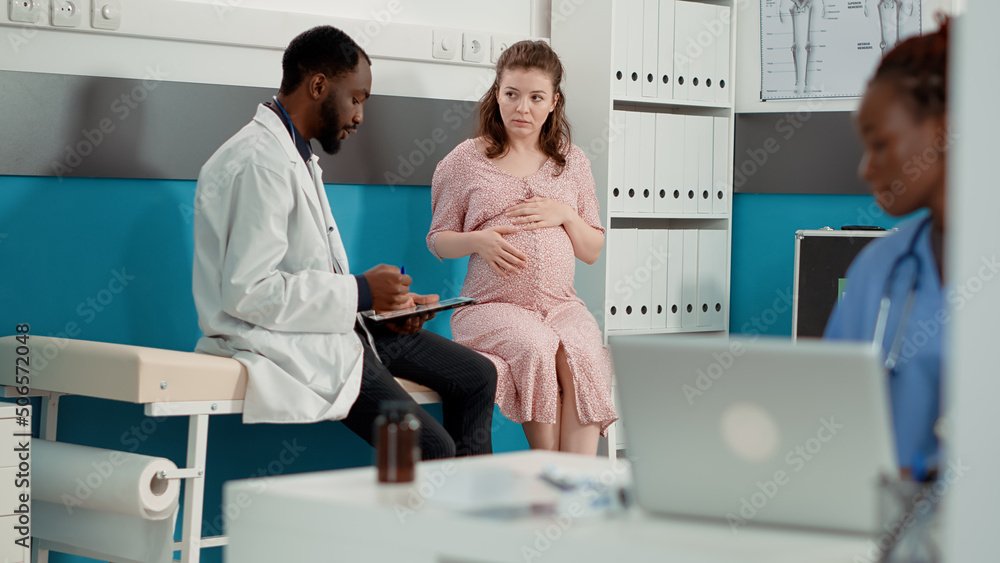 Obraz premium Health specialist doing checkup consultation with expectant patient in office. Male doctor using digital tablet to take notes, giving medical advice to woman with pregnancy belly.