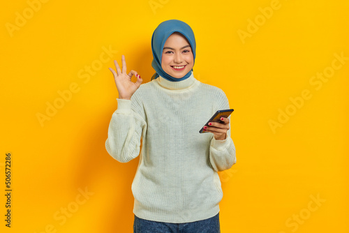 Portrait of smiling young asian woman in white sweater and hijab holding mobile phone, showing ok sign isolated over yellow background. People islam religious concept