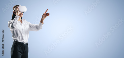 Photos Young woman working in virtual reality environment in VR glasses, presses by her finger on virtual interface on abstract blue backdrop