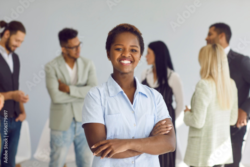 Portrait of happy successful confident young black businesswoman. Charming woman and business leader in smart casual shirt looking at camera and smiling standing in office with her team in background