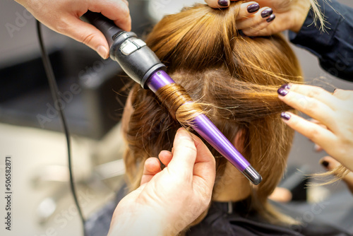 The hairstylist makes curls hairstyle of long brown hair with the curling iron in hairdresser salon, close up