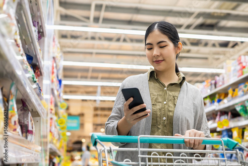 Asian woman checking shopping list on her smartphone in supermarket with trolley cart. Female using smartphone and application check product information in supply modern trade hall. photo