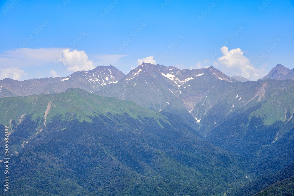 Fototapeta premium Mountain bright landscape with snow-capped mountains, blue sky and clouds. Mountainside covered with green grass and fir trees.