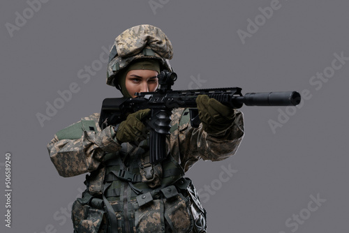 Portrait of militant woman dressed in camouflage clothes aiming rifle against gray background.