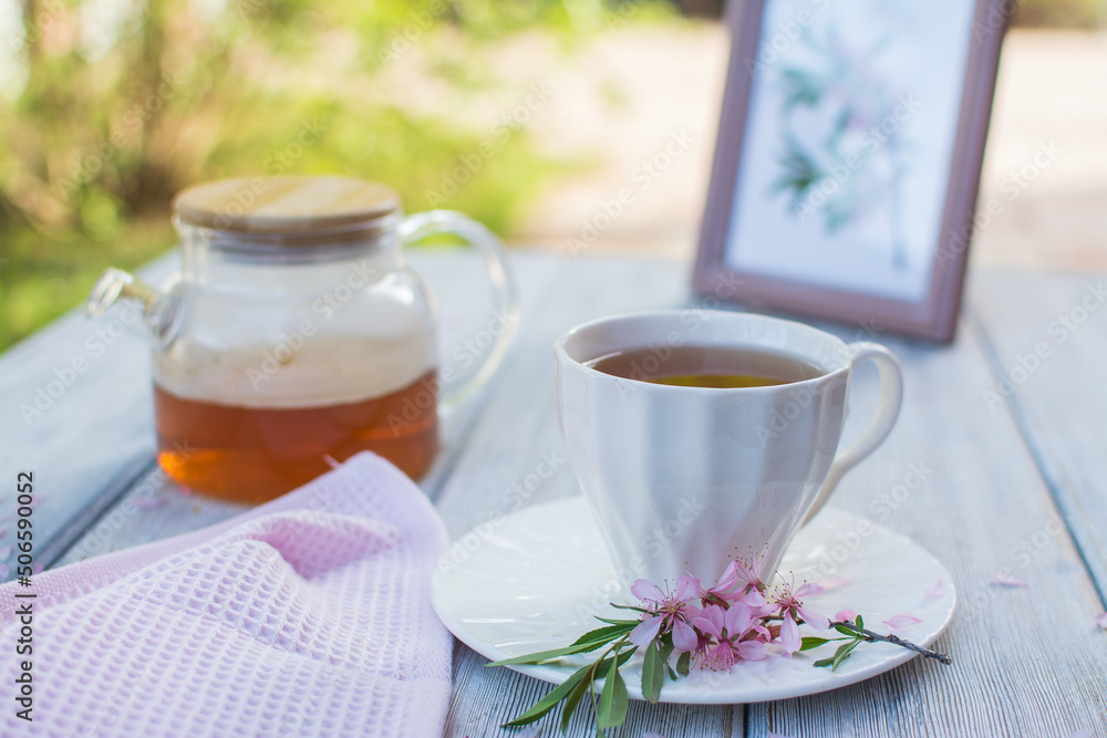 Glass teapot with tea and a white ceramic cup on a wooden table. In the background are flowers in a white jug and a painting. Glass teapot with tea and a white ceramic cup on a wooden table. In the background are flowers in a white jug and a painting.