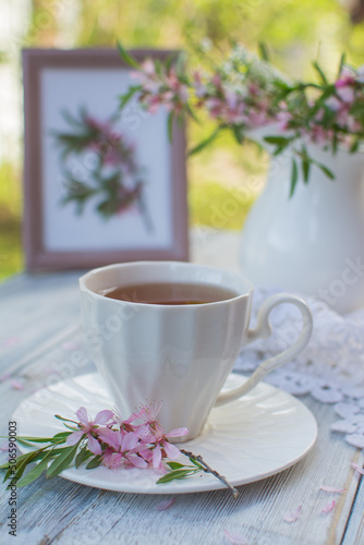 Cup with tea in a white ceramic cup on a wooden table. In the background are flowers in a white jug and a painting.