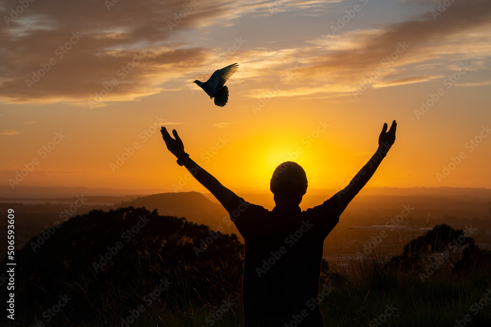 Silhouette of a man with open arms greeting the sun, bird flying away at sunrise. Hope concept.