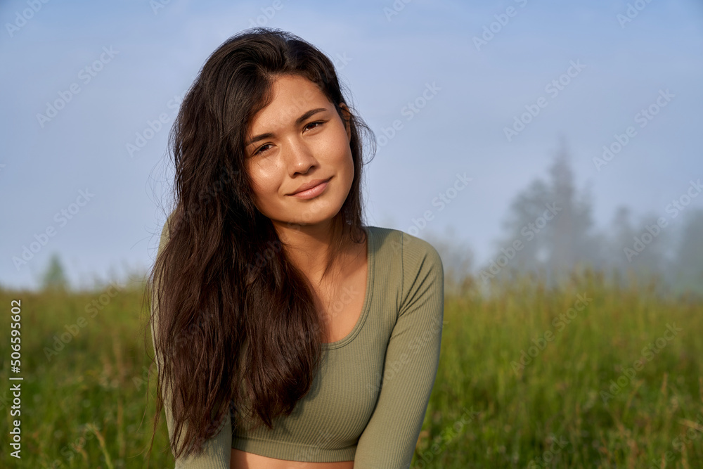 Fototapeta premium Portrait of young woman with dark hair sitting among green summer nature and looking at camera. Recreation during sport activity of pretty lady.