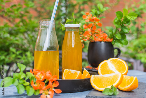 Two bottles of orange juice on a black ceramic plate. Near slices of an orange and a branch with flowers.