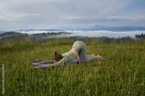 Active dark haired woman in sport clothes doing flexible exercises among green summer mountains. Sporty lady enjoying outdoors workout during free time. 