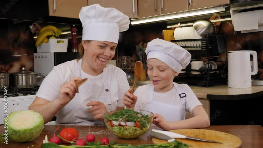 Sweet kind mother teaches her little curious son how to cook, they mix vegetable salad together with wooden spoons. Family in aprons and hats in the kitchen. Sweet kind mother teaches her little curious son how to cook, they mix vegetable salad together with wooden spoons. Family in aprons and hats in the kitchen.