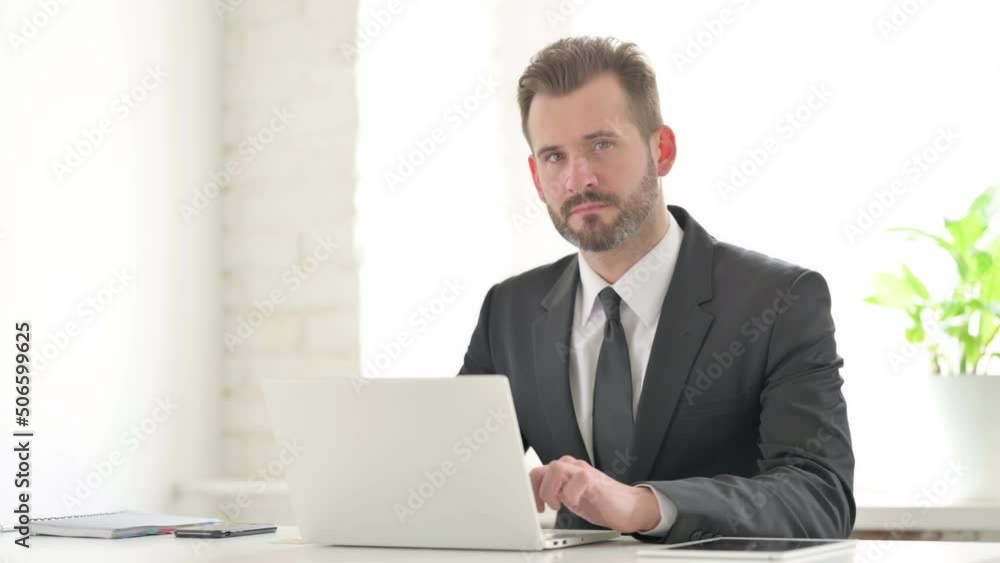Young Businessman Looking at Camera while using Laptop in Office Young Businessman Looking at Camera while using Laptop in Office