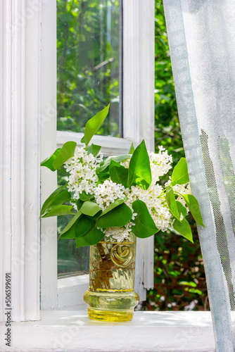 Fototapeta a bouquet of white lilac flowers in a glass vase on a white windowsill at an ope