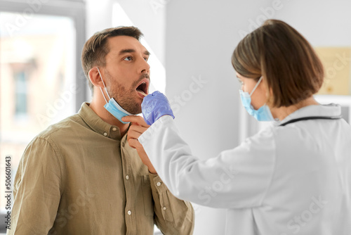 medicine, healthcare and people concept - female doctor in mask with tongue depressor checking sore throat of man patient at hospital