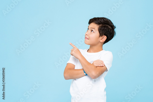 Cute smiling boy in plain white t shirt looking and pointing hand up in isolated studio light blue color background