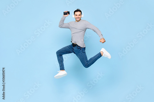 Cheerful young Caucasian man wearing headphones listening to music from smartphone and jumping in studio light blue color isolated background