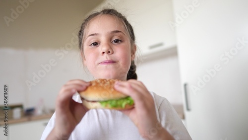 little kid girl eating a hamburger. unhealthy fast food meal proper nutrition concept. child greedily with pleasure bites a big burger in the kitchen at lifestyle home. kid eats fast food close-up