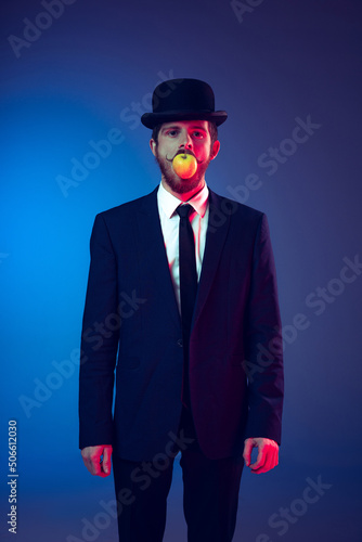 Creative portrait of young stylish man in business suit and bowler hat on his head isolated on dark blue studio background. Concept of art, fashion, theater, fun