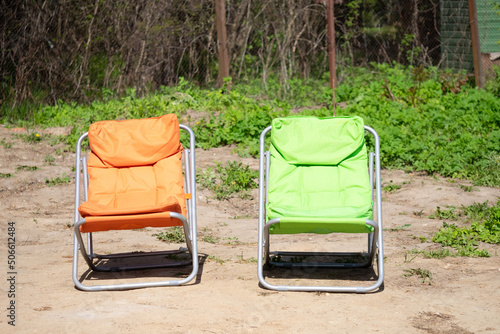 Orange and green folding tourist chair for leisure on the background of brown earth, green grass, iron mesh fence under sun