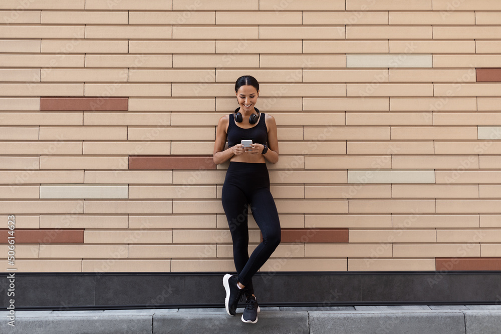 Smiling young slim muslim female in sports uniform with fitness tracker and headphones typing on smartphone Smiling young slim muslim female in sports uniform with fitness tracker and headphones typing on smartphone