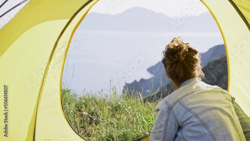 Lady traveler opens entrance in yellow tent. Young woman relaxing at nature enjoying beautiful view from hill top over green meadow and sea coast in sunny day