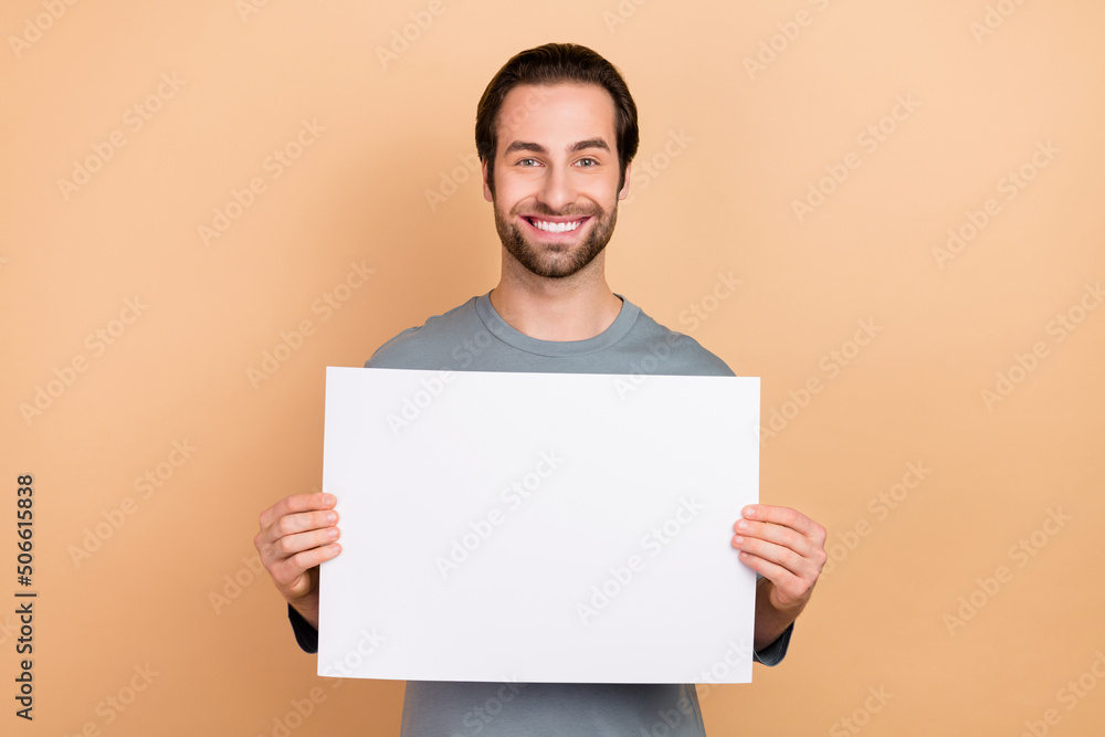 Photo of young cheerful man hold hands paper poster promo offer isolated over beige color background Photo of young cheerful man hold hands paper poster promo offer isolated over beige color background