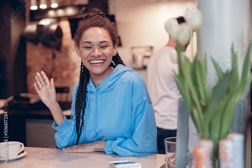 Pretty mixed-race young female bartender with piercing smiling at counter and waving hand. Portrait