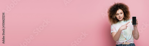 Young woman in t-shirt pointing at cellphone with blank screen isolated on pink, banner.