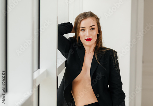 sexy beautiful girl in a black business suit with red lipstick on her lips and naked body. near the window. mesmerizing view