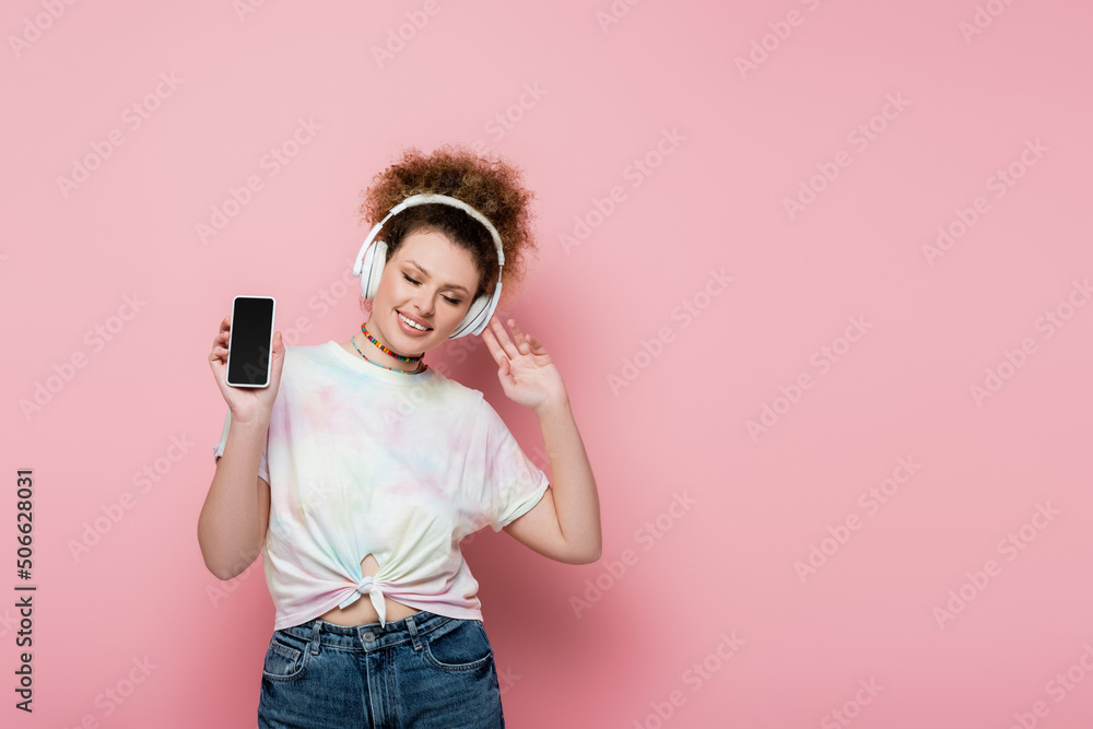 Curly woman in headphones holding cellphone on pink background. Curly woman in headphones holding cellphone on pink background.