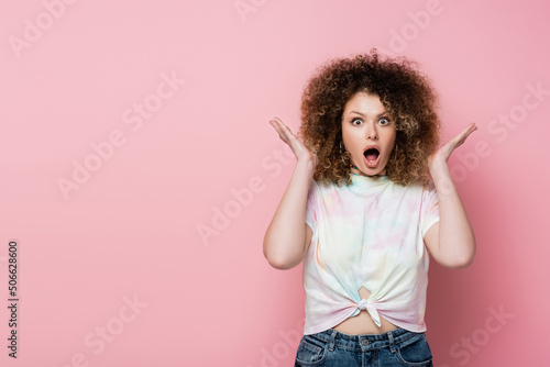 Astonished woman in t-shirt looking at camera on pink background.