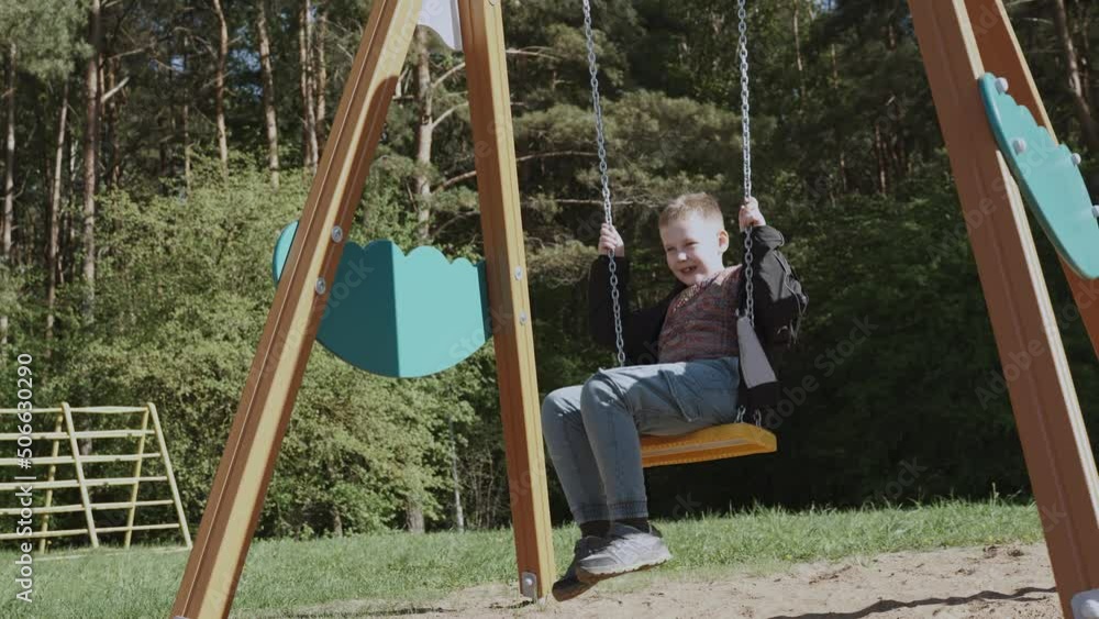 Happy boy schoolboy swings on wooden swing at the playground near the green forest on a sunny day. Child smiles and enjoys the rest. Swing on metal chains over the sand, slow motion. Happy boy schoolboy swings on wooden swing at the playground near the green forest on a sunny day. Child smiles and enjoys the rest. Swing on metal chains over the sand, slow motion.