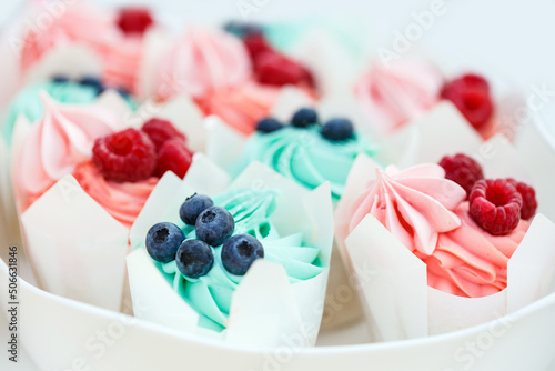 Appetizing and delicious muffins with pink and blue cream cheese are decorated with fresh wild raspberries and blueberries. Sweet tasty desserts on white plate. Boy or girl? Close-up, white background