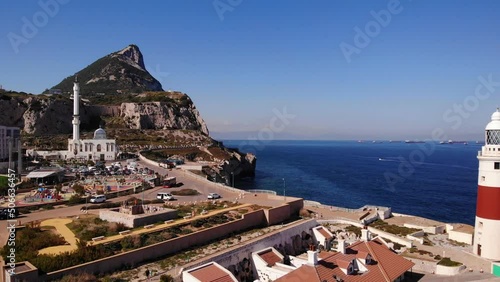 High tower of the White Ibrahim-al-Ibrahim Mosque and the Europa point lighthouse with the Rock of Gibraltar in the background with a view on cargo ships waiting at the anchorage. Drone trucking shot