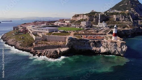 Strong swell creates a heady sea on a windless summer day as white gulls fly around the Europa point lighthouse in Gibraltar with in the background ships at the anchorage. Aerial drone lifting shot